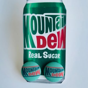 May include: A green Mountain Dew soda can with the words "Mountain Dew" in white and red. Below the can are two round, teal-colored buttons with the Mountain Dew logo.