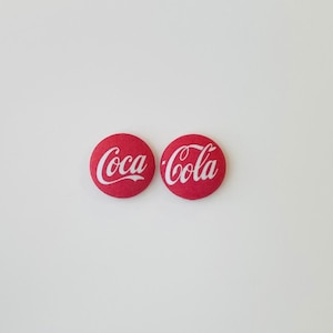 May include: Two red Coca-Cola button pins with white script lettering. The pins are round and have a fabric-like texture. The Coca-Cola logo is prominently displayed on each pin, a classic design.