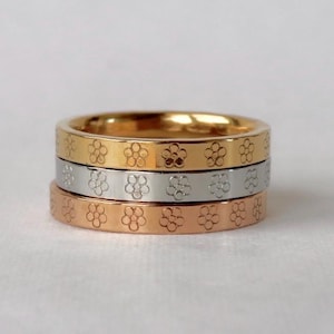 May include: Three stacked rings in gold, silver, and rose gold. Each ring features a repeating floral pattern around the band. The rings are displayed on a white surface.
