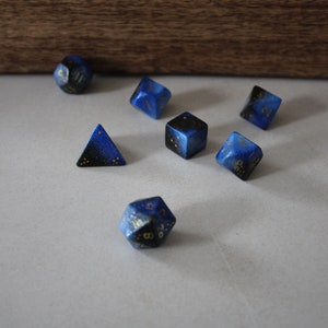 Puede incluir: Un conjunto de seis dados azules y negros con motas doradas. Los dados son de varias formas, incluyendo una pirámide, un cubo y un d20.