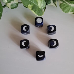 May include: Six black dice with white crescent moon and full moon designs. The dice are speckled with white glitter.