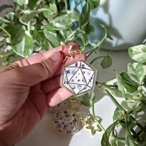 Può includere: Un portachiavi geometrico trasparente con chiusura a forma di stella dorata. Il portachiavi presenta un design floreale bianco e blu all'interno di una forma multi-lato bordata d'oro. Piante verdi sullo sfondo.