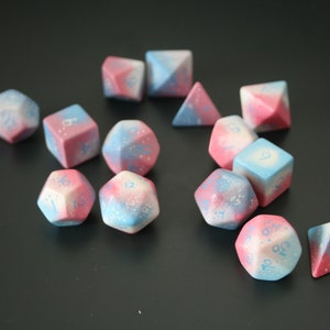 Può includere: Un set di dadi a 12 facce con un designo a galassia rosa e blu. I dadi sono fatti di un materiale maculato con numeri bianchi.