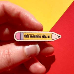 Op de afbeelding: Een houten speld in de vorm van een potlood met de tekst "this machine kills ai." Het potlood heeft een gele behuizing, een roze gum en een grijze ferrule. De achtergrond is geel en rood.