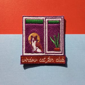 May include: Embroidered patch depicting a cat in a window, with a golden halo. The window is purple with green trim, and a small plant is visible. The text "window cat fan club" is below the window.