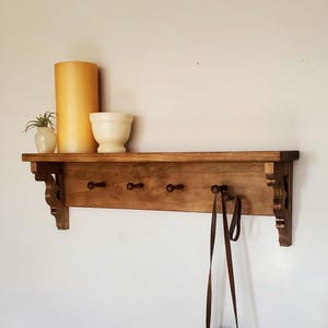 May include: A wooden wall shelf with a shelf, four pegs, and decorative corbels. A small leather bag hangs from one of the pegs. The shelf holds a candle, a small plant, and a white bowl. The shelf is mounted on a white wall.