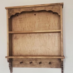 Puede incluir: Una estanter&iacute;a de pared de madera con acabado natural. Cuenta con una parte superior festoneada, dos estantes y una fila inferior de tres clavijas. La estanter&iacute;a est&aacute; montada en una pared blanca.