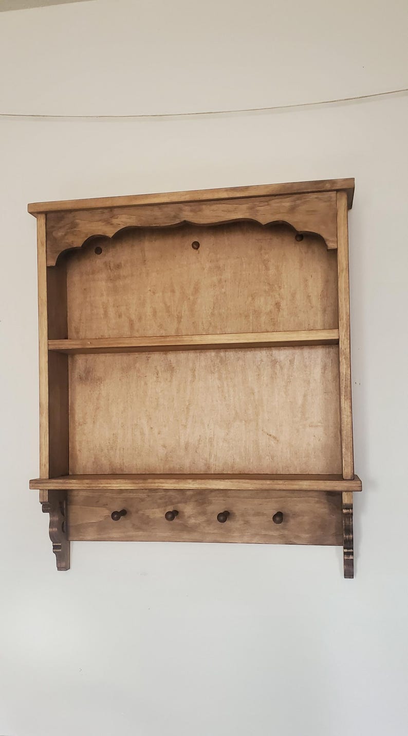 Puede incluir: Una estanter&iacute;a de pared de madera con acabado natural. Cuenta con una parte superior festoneada, dos estantes y una fila inferior de tres clavijas. La estanter&iacute;a est&aacute; montada en una pared blanca.