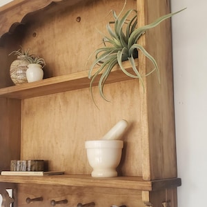 Puede incluir: Estanter&iacute;a de pared de madera con acabado natural, con m&uacute;ltiples estantes y una fila de clavijas. La estanter&iacute;a exhibe un mortero y una maja, plantas decorativas y una taza colgante. El dise&ntilde;o incluye soportes decorativos.