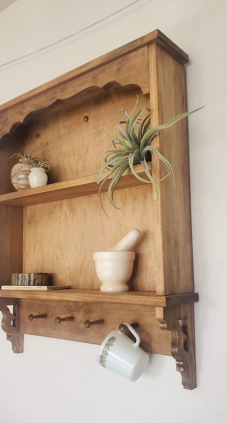 Puede incluir: Estanter&iacute;a de pared de madera con acabado natural, con m&uacute;ltiples estantes y una fila de clavijas. La estanter&iacute;a exhibe un mortero y una maja, plantas decorativas y una taza colgante. El dise&ntilde;o incluye soportes decorativos.