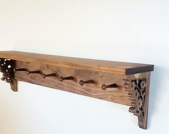 May include: A long, rectangular wooden wall shelf featuring a flat top surface and a series of small, protruding pegs for hanging items. The shelf is supported by two decorative, intricately carved wooden brackets on each side.
