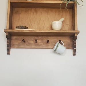 Puede incluir: Estanter&iacute;a de pared de madera con dos estantes, tres clavijas y soportes decorativos. La estanter&iacute;a contiene un jarr&oacute;n peque&ntilde;o, una planta, un mortero y una maja, y una taza con un dise&ntilde;o floral. La madera tiene un acabado c&aacute;lido y natural.
