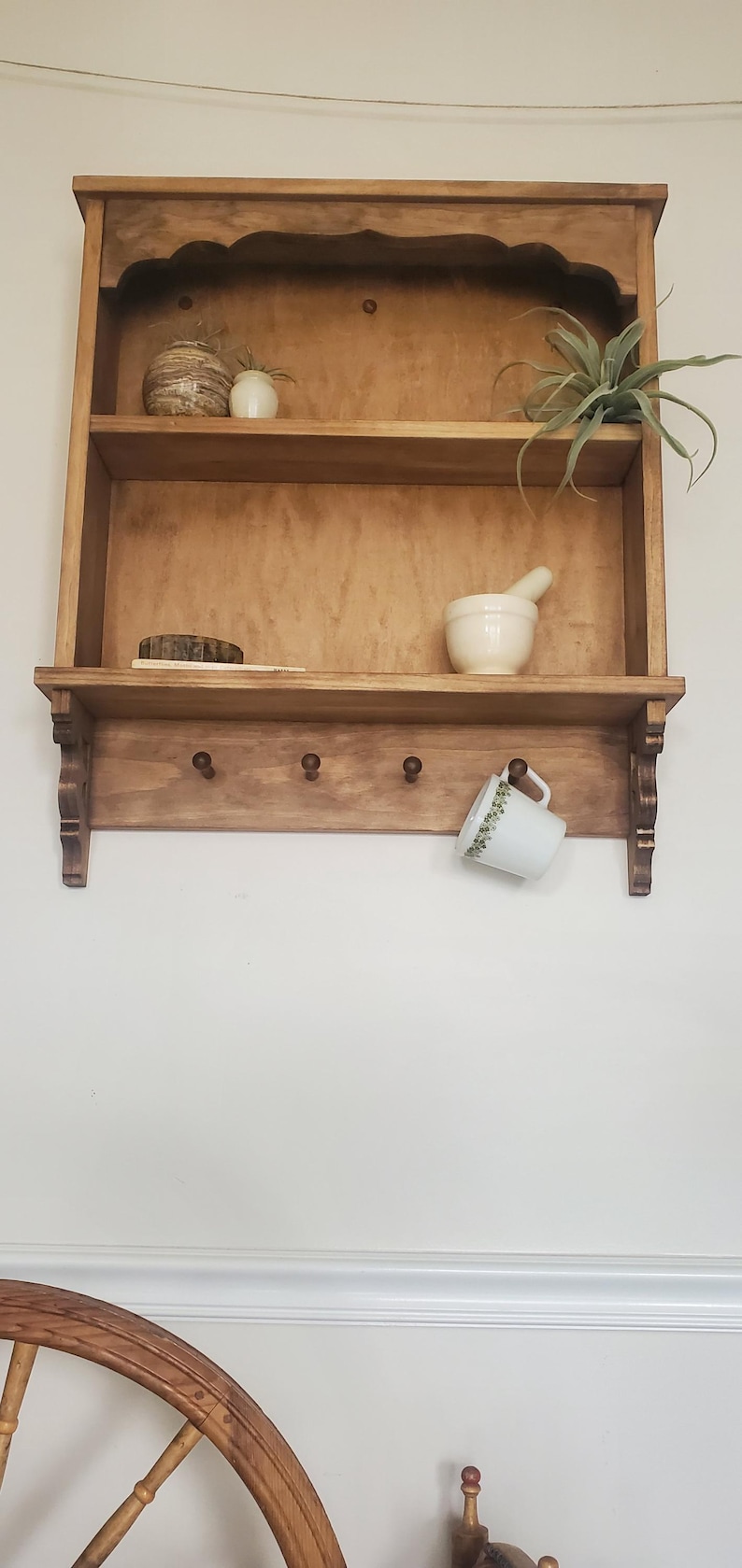 Puede incluir: Estanter&iacute;a de pared de madera con dos estantes, tres clavijas y soportes decorativos. La estanter&iacute;a contiene un jarr&oacute;n peque&ntilde;o, una planta, un mortero y una maja, y una taza con un dise&ntilde;o floral. La madera tiene un acabado c&aacute;lido y natural.
