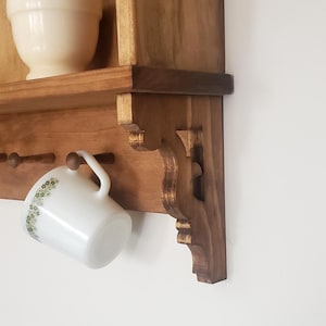 Puede incluir: Estante de pared de madera con un soporte decorativo. Una taza blanca con un estampado floral verde cuelga del estante. Un mortero y una maja blancos est&aacute;n en el estante.