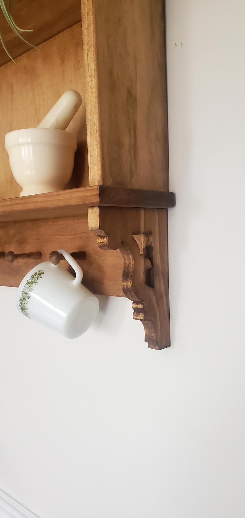 Puede incluir: Estante de pared de madera con un soporte decorativo. Una taza blanca con un estampado floral verde cuelga del estante. Un mortero y una maja blancos est&aacute;n en el estante.