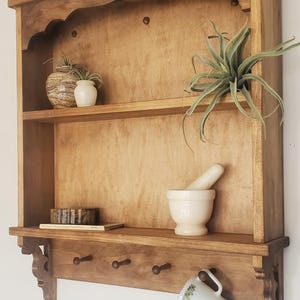 Puede incluir: Estanter&iacute;a de pared de madera con dos estantes y tres ganchos debajo. Exhibe peque&ntilde;as plantas en macetas, un mortero y una maja, y una taza. La madera tiene un acabado c&aacute;lido y natural.