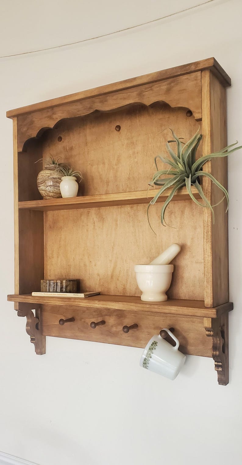 Puede incluir: Estanter&iacute;a de pared de madera con dos estantes y tres ganchos debajo. Exhibe peque&ntilde;as plantas en macetas, un mortero y una maja, y una taza. La madera tiene un acabado c&aacute;lido y natural.