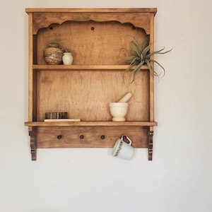 Puede incluir: Estanter&iacute;a de pared de madera marr&oacute;n claro con tres estantes y tres clavijas. Contiene un peque&ntilde;o jarr&oacute;n, un mortero y una taza. Una planta con hojas verdes largas est&aacute; en el estante superior.