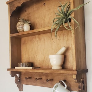 Puede incluir: Estante de pared de madera con dos estantes, un estante inferior con clavijas y soportes decorativos. El estante contiene un mortero y una maja, peque&ntilde;as plantas y una taza con estampado floral.