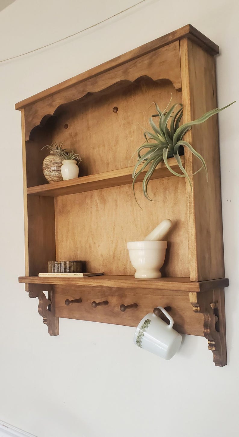 Puede incluir: Estante de pared de madera con dos estantes, un estante inferior con clavijas y soportes decorativos. El estante contiene un mortero y una maja, peque&ntilde;as plantas y una taza con estampado floral.