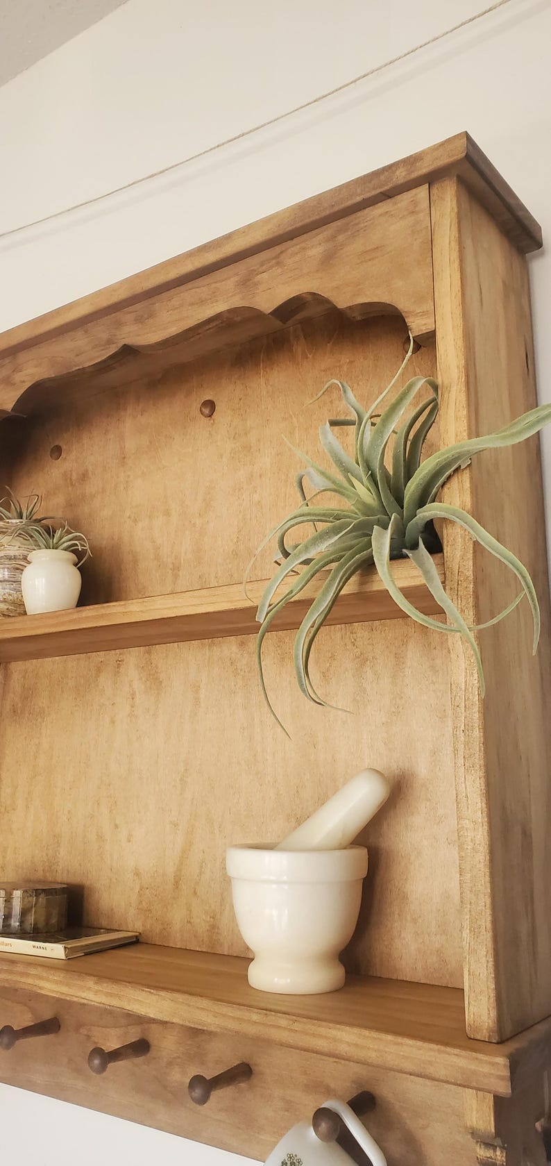 Puede incluir: Gabinete de madera con una parte superior festoneada, con un estante con un mortero y una maja blancos, una peque&ntilde;a planta en maceta y una planta a&eacute;rea. El gabinete tiene un acabado de madera natural e incluye una fila de clavijas.