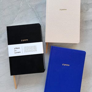 May include: Three hardcover journals with gold lettering. The journals are black, cream, and blue. The black journal has the text "5 lines for 5 years" on the front. The cream journal has the text "5 years" on the front. The blue journal has the text "5 years" on the front.