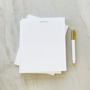 May include: A stack of white paper with the text "BRILLIANT IDEAS" printed on the top sheet. A white marker with a gold cap is lying on a grey surface next to the paper.