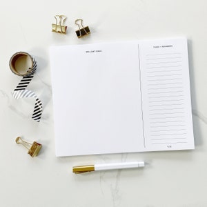 May include: A white notepad with a black and white striped washi tape, two gold binder clips, a gold paper clip, and a gold pen. The notepad is divided into two sections, one labelled "Brilliant Ideas" and the other labelled "Tasks + Reminders".