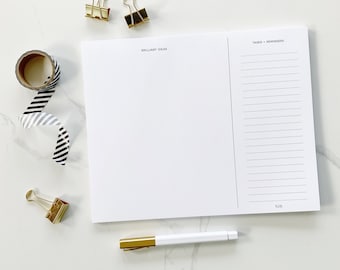 May include: A white notepad with a black and white striped washi tape, two gold binder clips, a gold paper clip, and a gold pen. The notepad is divided into two sections, one labeled "Brilliant Ideas" and the other labeled "Tasks + Reminders".