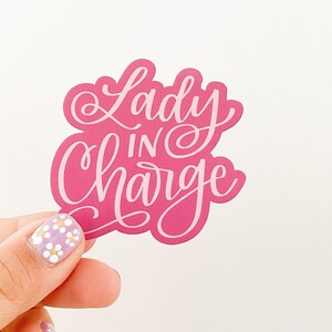 May include: Pink sticker with white lettering that reads "Lady in Charge".