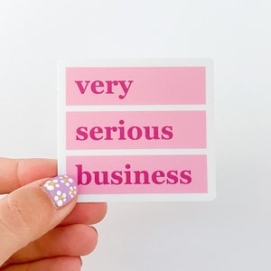 May include: A pink and white sticker with the words "very serious business" in dark pink. The text is arranged in three horizontal lines. The sticker is held by a hand with a floral nail design.