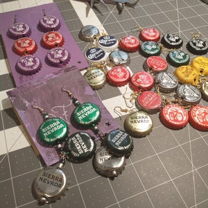 May include: Handmade earrings crafted from repurposed bottle caps, featuring various beer and soda brands. The earrings are displayed on a purple card and a gray cutting mat, with jewelry-making tools in the background.