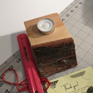 May include: A wooden candle holder with a natural bark finish and a small white tea light candle. The candle holder is sitting on a gray cutting mat with a red measuring tape.