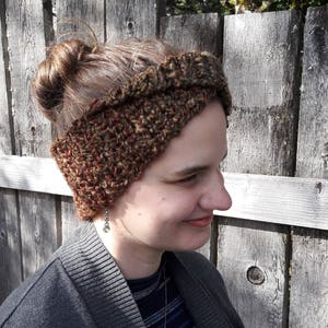 May include: A brown and green crocheted headband with a twisted design. The headband is worn by a person with brown hair and a grey cardigan.