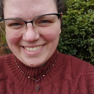 Può includere: Una collana di perline con un piccolo ciondolo a forma di pigna. La collana presenta perline marroni, dorate e viola. La persona nella foto indossa un maglione rosso.