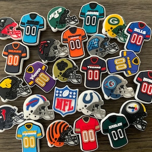 Puede incluir: Una colección de coloridos dijes para zapatos con logotipos y camisetas de equipos de fútbol americano. Los dijes incluyen cascos y camisetas con nombres y números de equipos, como los Jets, Jaguars y Bills.