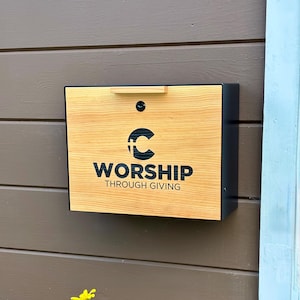 May include: A rectangular wall-mounted donation box with a light wood grain front panel and black metal sides. The front features a black logo of a letter C with a cross and the text WORSHIP THROUGH GIVING. It includes a small handle and lock.