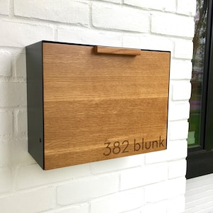 May include: A rectangular mailbox with a wooden front and a black metal frame is mounted on a white brick wall. The wooden front has a natural wood grain and a small handle. The text "382 blunk" is etched on the front.