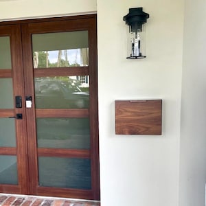 May include: A wooden mailbox with a natural finish is mounted on a white wall, next to a black outdoor light fixture. The mailbox has a rectangular shape with a small handle. A wooden door with glass panels is on the left.