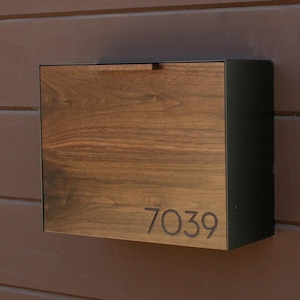Könnte beinhalten: Ein rechteckiger Wandbriefkasten mit einer Frontplatte aus dunkelbraunem Holzmaserung und einem horizontalen Griff an der Oberseite. Die Zahl 7039 ist in die untere rechte Ecke der Holzplatte eingraviert, die Seiten sind mattschwarz.