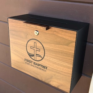 Può includere: Una cassetta di raccolta rettangolare da parete con telaio in metallo nero opaco e pannello frontale in legno naturale. Il pannello in legno è inciso con un emblema a croce circolare e il testo FIRST BAPTIST FORT WALTON BEACH. Include una serratura.