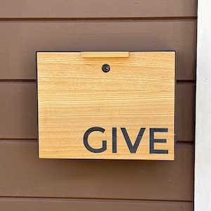 Puede incluir: Una caja de donación de madera con un acabado de veta de madera clara. La caja tiene un borde negro y una tapa con bisagras con un pequeño asa. La palabra "GIVE" está impresa en letras negras en negrita en la parte delantera.