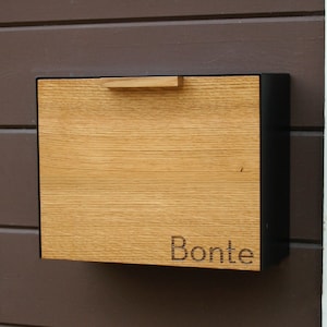May include: A wooden mailbox with a black metal frame. The mailbox has a wooden door with a handle and the name "Bonté" engraved on the front.