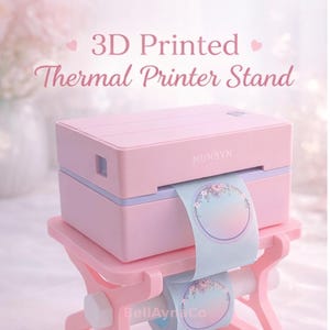 May include: A pink 3D printed thermal printer stand with a matching pink thermal printer. The printer is labeled "MUNBYN" and is dispensing a roll of light blue stickers with a floral design. The stand is also pink and has a delicate, decorative design.