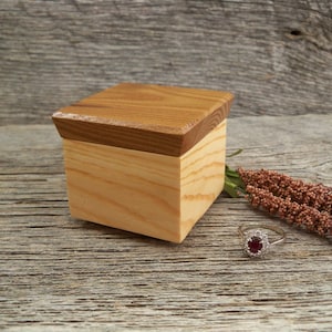 May include: A small wooden box with a hinged lid, made of light brown wood. The box is sitting on a rustic wooden surface, next to a red gemstone ring and a dried flower sprig.