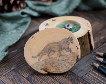 May include: A wooden ring box with a cougar illustration on the lid. The box is made from a cross-section of a tree branch, with a natural wood grain finish. The box is open, revealing a ring inside.