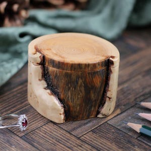 May include: A small, handcrafted wooden box with a natural, light-colored exterior and a dark, textured interior. A silver ring with a red gemstone sits nearby. The box is on a wooden surface.