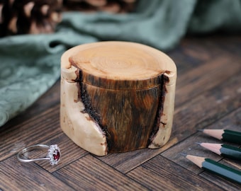 May include: A small, handcrafted wooden box with a natural, light-colored exterior and a dark, textured interior. A silver ring with a red gemstone sits nearby. The box is on a wooden surface.