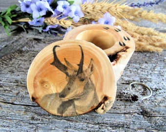 May include: A wooden ring box with a burnt wood design of a pronghorn antelope on the lid. The box is open and the inside is empty. The box is made of natural wood and has a rustic look.