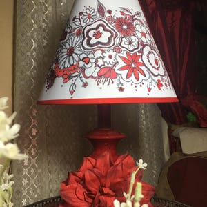 May include: A red table lamp with a white lampshade featuring a floral design in red and black. The lamp base is red, and a large red flower sits at the base. The lamp is on a dark brown tray.